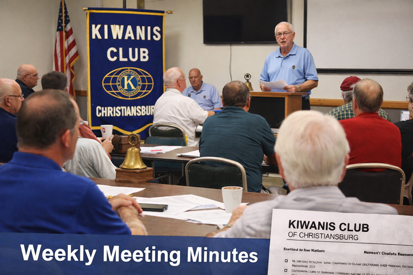 Photorealistic image of a Kiwanis Club of Christiansburg weekly meeting in a community room. Members sit at tables reviewing printed agendas while the club president stands at a podium speaking. A Kiwanis banner and American flag are displayed behind him. A blue banner across the bottom of the image reads “Weekly Meeting Minutes.”